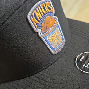 Op de afbeelding: Een zwarte honkbalpet met een geperforeerd zijpaneel en een geborduurde patch aan de voorzijde. De patch toont een basketbalafbeelding met de tekst KNICKS in oranje en blauw. Een ronde zwarte sticker is bevestigd op de platte klep van de pet.