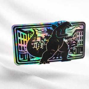 May include: A black Godzilla-themed sticker with a holographic border. The sticker features a silhouette of Godzilla destroying a city. The background shows buildings and a cityscape. The sticker is rectangular.