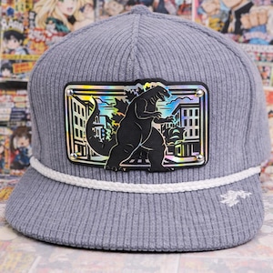 May include: Gray corduroy baseball cap with a white rope detail and a Godzilla patch. The patch features a black Godzilla silhouette against a cityscape with iridescent accents. The cap has a flat brim and a small embroidered design on the side.