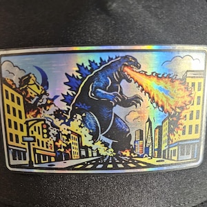 Op de afbeelding: Een rechthoekige patch met een holografische afbeelding van Godzilla die een stad vernietigt. Het monster is donkerblauw en spuwt vuur. Gebouwen staan in brand, met een maan in de lucht.