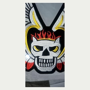 May include: Embroidered patch featuring a skull design with flames, wings, and a black background. The skull is white with yellow eyes and a wide grin. The flames are red and yellow, and the wings are yellow and white.