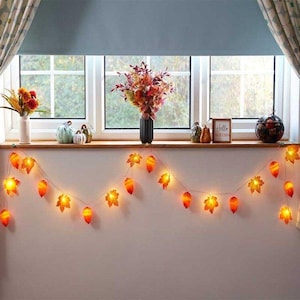 May include: A cosy autumn display with a string of leaf-shaped fairy lights. The lights are orange and yellow, strung below a window. The window sill features vases of flowers, pumpkins, and a sign that reads "Hello Autumn".