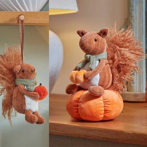 May include: A plush squirrel ornament with a cord for hanging and a matching plush squirrel figurine sitting on a pumpkin. Both squirrels are brown with white bellies, fluffy tails, and green scarves. The hanging squirrel holds a small orange pumpkin.
