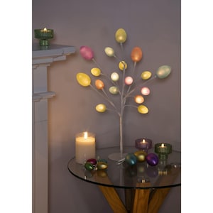 May include: An Easter tree with egg-shaped lights in pastel colours. The tree is on a round glass table with lit candles and decorative eggs. A green glass candle holder is on a white surface.