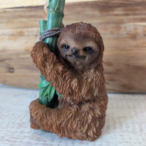 May include: A brown sloth ornament holding onto a green bamboo stalk. The sloth has detailed fur and a smiling face. The ornament is a decorative item.