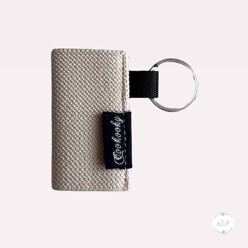 Peut inclure: Un porte-cl&eacute;s en tissu beige avec une &eacute;tiquette noire sur laquelle est inscrit "Cuckoocky" et un anneau de cl&eacute; argent&eacute;.