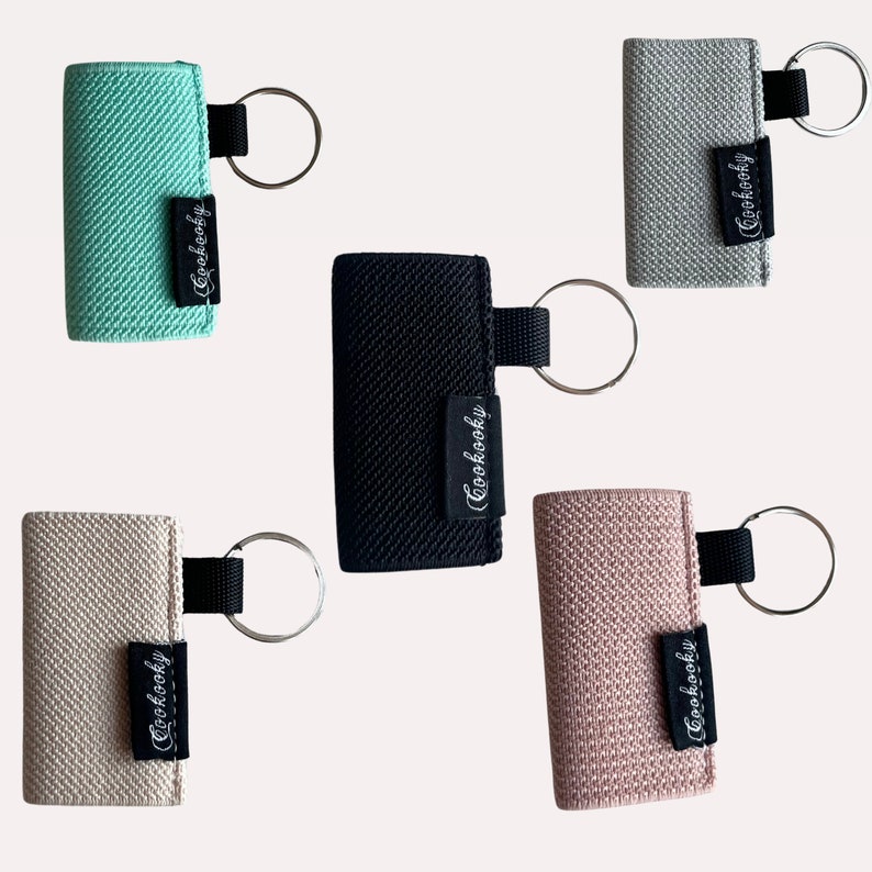 Peut inclure: Cinq porte-cl&eacute;s avec des housses en tissu de diff&eacute;rentes couleurs et un anneau argent&eacute;. Les housses en tissu sont turquoise, noir, beige, rose et gris. Chaque porte-cl&eacute;s porte l'inscription "Cuckooby" imprim&eacute;e sur la housse en tissu.