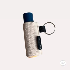 Peut inclure: Un porte-cl&eacute;s en tissu beige avec un tube de baume &agrave; l&egrave;vres bleu et jaune &agrave; l'int&eacute;rieur. Le porte-cl&eacute;s a une sangle noire et un anneau argent&eacute;.