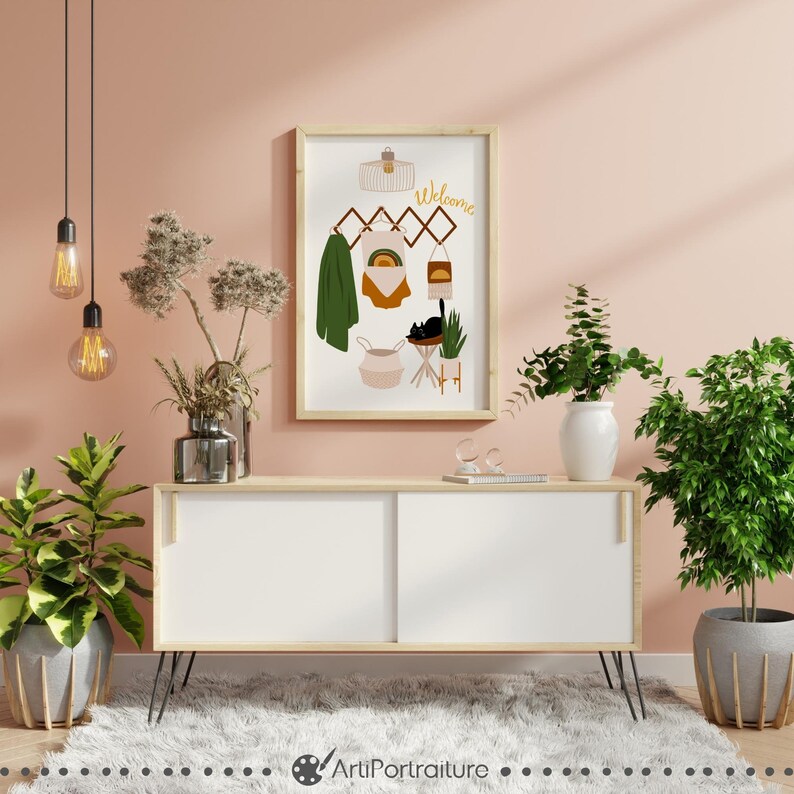 May include: A framed illustration of a welcome sign with a rainbow, a cat, a basket, and a plant. The illustration is hanging on a wall with a light pink background.