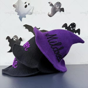 May include: Two knitted witch hats, one black and one purple, are stacked. The purple hat has the name "Mildred" embroidered in black. The black hat has "Addie" embroidered in purple. The background features Halloween-themed paper cutouts.