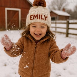 Op de afbeelding: Een kind draagt een crèmekleurige gebreide muts met de naam "EMILY" in bruine letters, een bruine pompon, een bruin fleecejack en roze wanten, spelend in de sneeuw. Een rode schuur staat op de achtergrond.