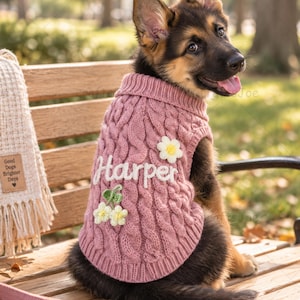 Puede incluir: Un su&eacute;ter de punto trenzado rosa para perro con el nombre Harper en letras cursivas blancas, decorado con peque&ntilde;as aplicaciones de flores blancas y amarillas. El su&eacute;ter tiene cuello y dobladillo acanalados, usado por un cachorro de pastor alem&aacute;n.