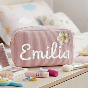 Puede incluir: Un neceser de maquillaje de pana rosa con el nombre "Emilia" bordado en escritura blanca y una margarita blanca. Artículos de maquillaje, incluyendo un pincel, pintalabios y un espejo, están dispuestos sobre una superficie de madera.