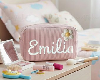 May include: A pink corduroy cosmetic bag with the name "Emilia" embroidered in white script, and a white daisy. Makeup items, including a brush, lipsticks, and a mirror, are arranged on a wooden surface.