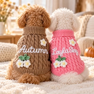 Puede incluir: Dos perros con suéteres de punto trenzado. El perro marrón lleva un suéter marrón con el texto Autumn y aplicaciones de flores blancas. El perro blanco lleva un suéter rosa con el texto Layla Sky y aplicaciones de flores blancas.