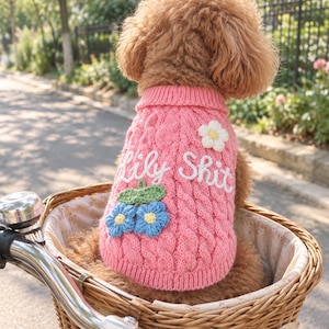 May include: A pink cable-knit dog jumper featuring the embroidered white text "Lily Shit" across the back. The garment is decorated with a white crocheted flower and two blue crocheted flowers with green leaves attached to the knit fabric.