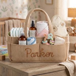 Könnte beinhalten: Eine beige Baby-Organizer-Tasche aus Cord mit dem Namen "Parker" auf der Vorderseite. Die Tasche enthält Windeln, Baby-Lotion, eine bärenförmige Decke, eine Rassel und andere Baby-Utensilien. Die Tasche hat einen Griff zum einfachen Tragen.