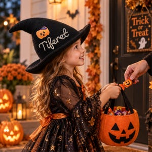 May include: A black knit witch hat featuring a small orange pumpkin patch and the embroidered name Tiferet in white script. The hat is worn with a black dress adorned with gold stars and moons, paired with an orange felt pumpkin candy bucket.