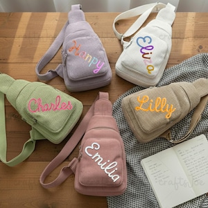 May include: A collection of corduroy sling bags in various colors, including green, lavender, pink, beige, and white. Each bag features a name embroidered on the front in a contrasting color. The bags have adjustable straps and a small front pocket.