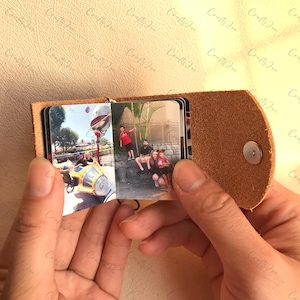 以下が含まれることがあります： 金属製の留め具が付いた茶色の革製のフォトアルバム。アルバムが開いていて、2枚の写真が見えます。最初の写真は、遊園地にいる4人家族の写真です。2枚目の写真は、ヤシの木の前に立っている4人家族の写真です。