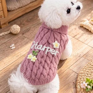 Puede incluir: Un perro peque&ntilde;o de color blanco lleva puesto un su&eacute;ter de punto trenzado en color rosa empolvado. El su&eacute;ter tiene el nombre Harper bordado en letra cursiva blanca, decorado con peque&ntilde;as flores tridimensionales blancas y amarillas.