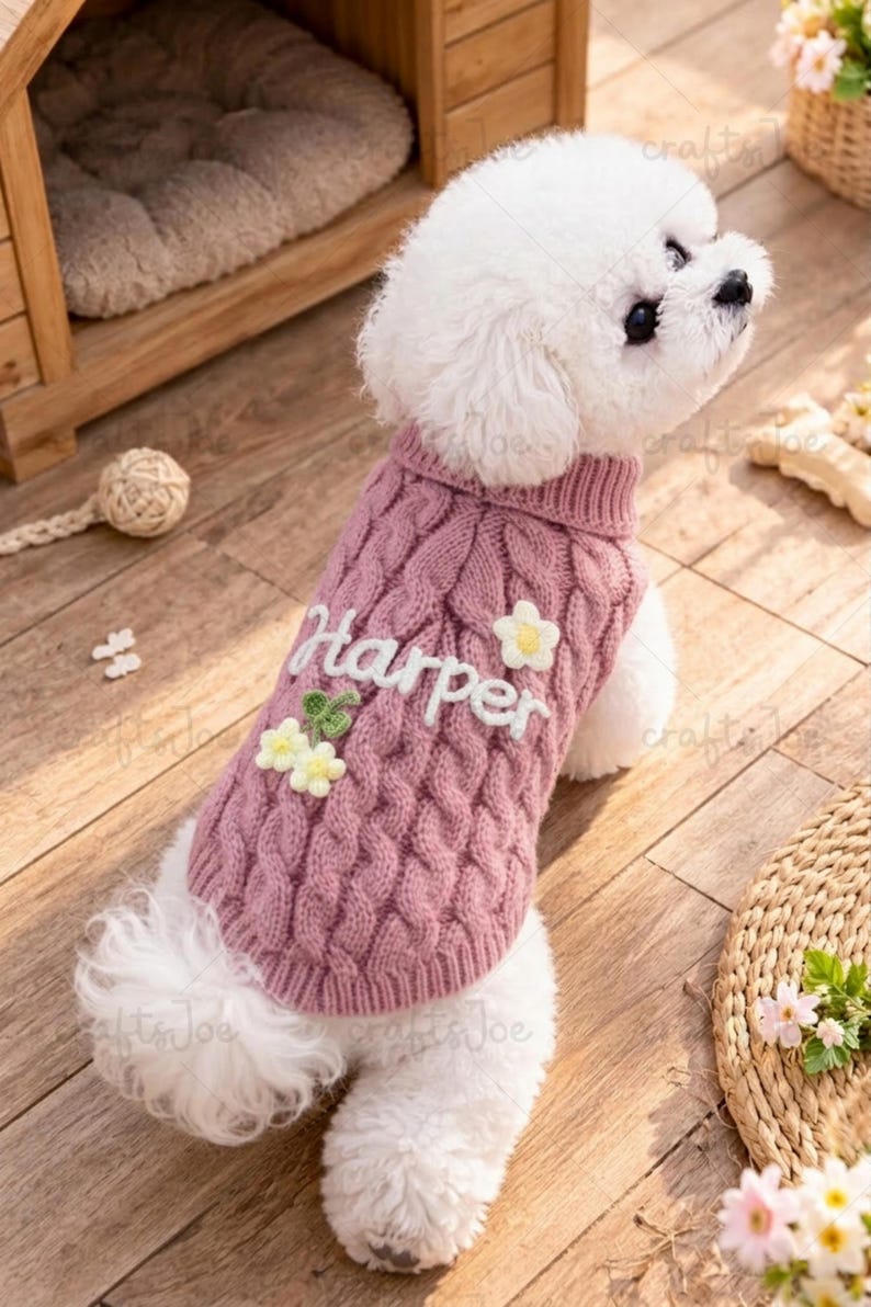Puede incluir: Un perro peque&ntilde;o de color blanco lleva puesto un su&eacute;ter de punto trenzado en color rosa empolvado. El su&eacute;ter tiene el nombre Harper bordado en letra cursiva blanca, decorado con peque&ntilde;as flores tridimensionales blancas y amarillas.