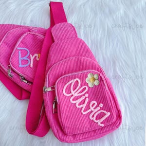 Könnte beinhalten: Hellrosa Cord-Umhängetasche mit einer Vordertasche und einem Reißverschluss. Die Tasche trägt den Namen "Olivia" in weißer Schreibschrift und eine kleine Blume. Eine weitere Tasche mit dem Namen "Bri" ist ebenfalls sichtbar.