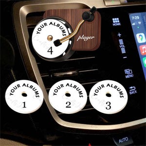 May include: A car air vent accessory with a miniature turntable design. The design includes a wooden base and a gold-coloured tone arm. The text "YOUR ALBUMS" is visible. Additional round discs with numbers 1-4 are also visible.