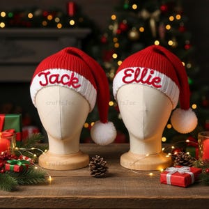 Puede incluir: Dos gorros de Papá Noel de punto rojo y blanco con pompones blancos en las puntas. El gorro izquierdo tiene bordado el nombre Jack en rojo, y el derecho el nombre Ellie en rojo, ambos sobre un borde blanco de canalé.