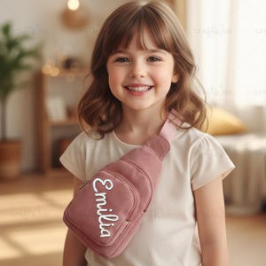 May include: A dusty rose corduroy sling bag with the name "Emilia" in white script. The bag has a zippered main compartment and a smaller front pocket. The bag is worn across the chest.