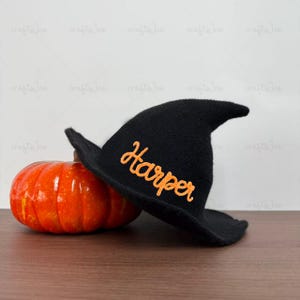 May include: A black knit witch hat featuring the name Harper embroidered in bright orange cursive script, positioned next to a glossy, vibrant orange pumpkin with deep vertical ridges and a smooth, reflective surface finish.