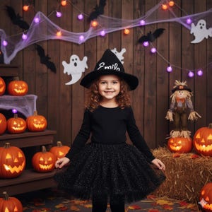 May include: A black witch hat with the name Emila embroidered in white script, paired with a long-sleeved black shirt and a black glittery tulle tutu skirt. The ensemble is designed for a Halloween costume, featuring a pointed hat structure.