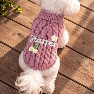 Puede incluir: Un su&eacute;ter para perro de punto trenzado en color rosa empolvado con cuello alto acanalado. La parte posterior del su&eacute;ter tiene bordado el texto blanco "Harper" y est&aacute; decorado con peque&ntilde;as flores aplicadas en relieve.