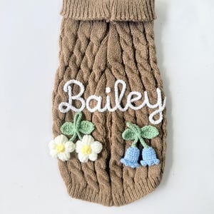 Puede incluir: Un su&eacute;ter de punto trenzado color canela con el nombre Bailey bordado en blanco. El su&eacute;ter est&aacute; decorado con dos flores blancas de ganchillo con centros amarillos a la izquierda y dos flores azul claro en forma de campana a la derecha.