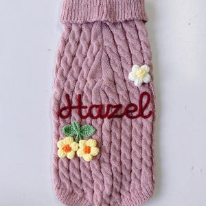 Puede incluir: Un su&eacute;ter para mascota de punto trenzado en color rosa empolvado con cuello alto acanalado. La prenda est&aacute; adornada con una flor de ganchillo blanca y dos flores de ganchillo amarillas con hojas verdes. El nombre Hazel est&aacute; bordado en rojo.