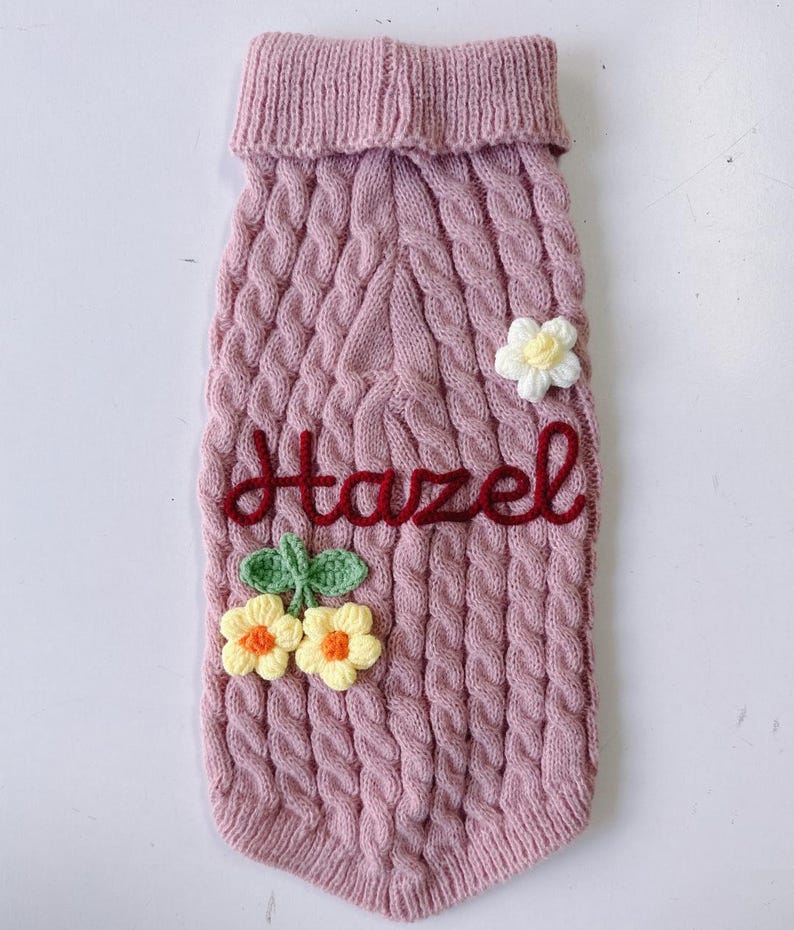 Puede incluir: Un su&eacute;ter para mascota de punto trenzado en color rosa empolvado con cuello alto acanalado. La prenda est&aacute; adornada con una flor de ganchillo blanca y dos flores de ganchillo amarillas con hojas verdes. El nombre Hazel est&aacute; bordado en rojo.