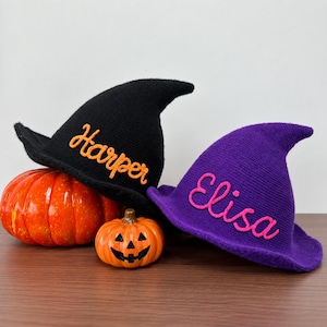 May include: A black knitted witch hat with the name Harper embroidered in orange and a purple knitted witch hat with the name Elisa embroidered in pink, positioned next to a large orange pumpkin and a small jack-o'-lantern with a carved face.