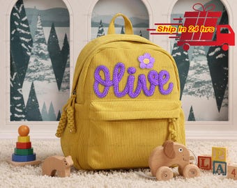 May include: A yellow corduroy backpack with the name "Olive" in purple script and a purple flower. The backpack has a front pocket and a top handle. Wooden toys, including a bear, elephant, and stacking rings, are in the foreground.