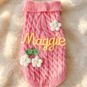 Puede incluir: Un su&eacute;ter de punto trenzado rosa para mascota con cuello y dobladillo acanalados. El su&eacute;ter est&aacute; decorado con la palabra Maggie escrita en hilo amarillo, una flor de ganchillo blanca y un par de flores de ganchillo blancas con hojas verdes.