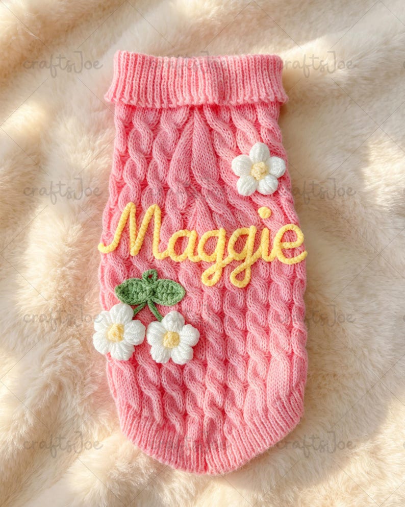 Puede incluir: Un su&eacute;ter de punto trenzado rosa para mascota con cuello y dobladillo acanalados. El su&eacute;ter est&aacute; decorado con la palabra Maggie escrita en hilo amarillo, una flor de ganchillo blanca y un par de flores de ganchillo blancas con hojas verdes.