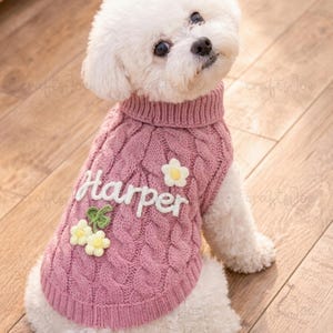 Puede incluir: Un perro blanco lleva puesto un su&eacute;ter de punto trenzado rosa polvoriento con cuello alto. El su&eacute;ter presenta el nombre Harper bordado en blanco, una aplicaci&oacute;n de flor blanca y un grupo de dos flores blancas con hojas verdes.