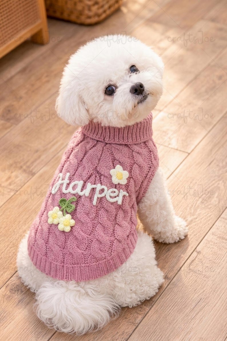 Puede incluir: Un perro blanco lleva puesto un su&eacute;ter de punto trenzado rosa polvoriento con cuello alto. El su&eacute;ter presenta el nombre Harper bordado en blanco, una aplicaci&oacute;n de flor blanca y un grupo de dos flores blancas con hojas verdes.