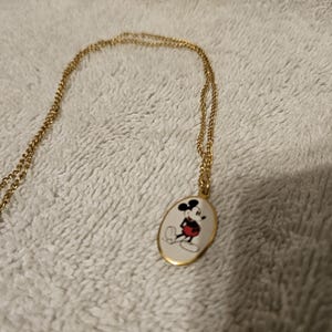 Puede incluir: Collar dorado con un colgante ovalado con una imagen de dibujos animados de Mickey Mouse. El colgante tiene un fondo blanco con un dise&ntilde;o de Mickey Mouse negro, rojo y amarillo. La cadena y el cierre tambi&eacute;n son dorados.