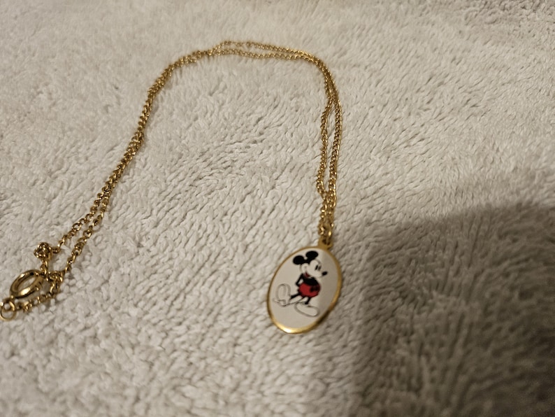 Puede incluir: Collar dorado con un colgante ovalado con una imagen de dibujos animados de Mickey Mouse. El colgante tiene un fondo blanco con un dise&ntilde;o de Mickey Mouse negro, rojo y amarillo. La cadena y el cierre tambi&eacute;n son dorados.