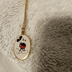 Puede incluir: Collar dorado con un colgante ovalado con Mickey Mouse. El colgante muestra a Mickey con sus pantalones cortos rojos, guantes blancos y silueta negra sobre un fondo blanco. La cadena es de dise&ntilde;o fino y enlazado.