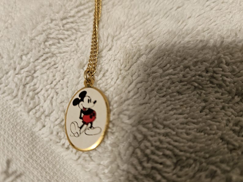 Puede incluir: Collar dorado con un colgante ovalado con Mickey Mouse. El colgante muestra a Mickey con sus pantalones cortos rojos, guantes blancos y silueta negra sobre un fondo blanco. La cadena es de dise&ntilde;o fino y enlazado.