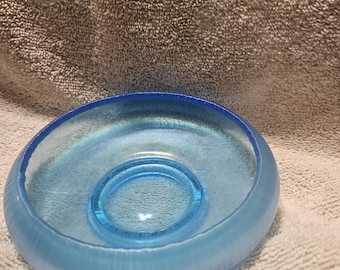 May include: A light blue glass bowl with a circular indentation in the center. The bowl has a rounded shape and a textured surface. The rim of the bowl is a darker shade of blue.