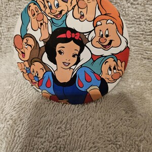 Puede incluir: Un botón redondo con Blancanieves y los Siete Enanitos. Blancanieves está en el centro, rodeada por los enanitos, todos en colores vibrantes. El botón tiene un fondo blanco y es probablemente un artículo de colección.