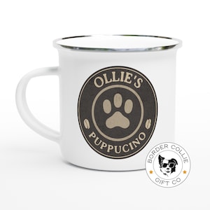 Puede incluir: Taza de esmalte blanco con un logotipo negro y marrón en forma de huella de pata que dice "Ollie's Puppuccino" en un círculo. La taza tiene un borde plateado. Debajo del logotipo hay un logotipo más pequeño que dice "Border Collie Gift Co."