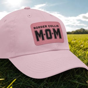 May include: A light pink baseball cap with a curved brim. The cap features a rectangular patch with the words "BORDER COLLIE MOM" in black block letters. The word "MOM" includes paw print details. The cap is made of a soft material.
